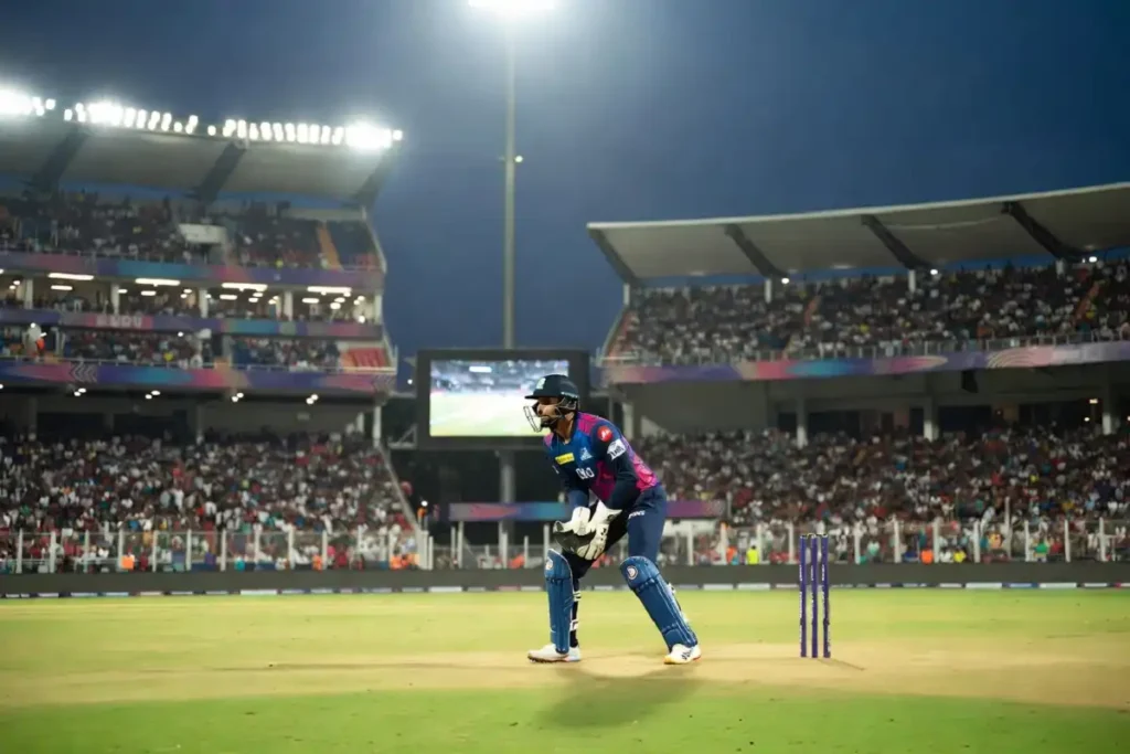 Cricket wicketkeeper in IPL jersey positioned behind the stumps in a floodlit stadium
